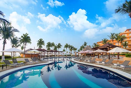 A luxurious resort pool with lounge chairs, umbrellas, and palm trees under a bright blue sky with scattered clouds; orange buildings are visible in the background—perfect for conference events and meetings at Mexico hotels.