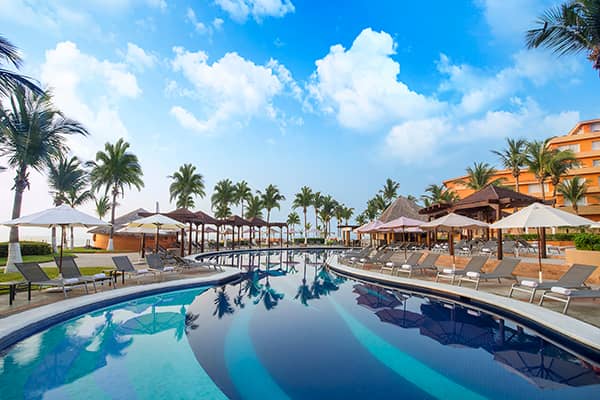 A tropical resort pool with lounge chairs, umbrellas, and palm trees on either side. The pool curves gently, reflecting the blue sky and clouds—perfect for conference events at Mexico hotels amid orange resort buildings and lush greenery.