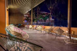 Terraza exterior por la noche con modernas sillas de mimbre, mesas redondas de cristal y cojines estampados. Al fondo se aprecian palmeras y un edificio iluminado con luz cálida, ideal para eventos y conferencias en hoteles de lujo en México.