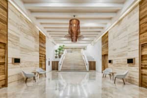 Amplio vestíbulo moderno con suelos de mármol pulido, paneles de madera en las paredes y sillas minimalistas, ideal para eventos y conferencias. Una singular lámpara de araña de madera cuelga de un techo con vigas a la vista, mientras que plantas en macetas adornan las escaleras de este elegante hotel resort en México.