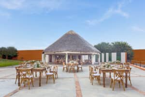Espacio para eventos al aire libre con mesas y sillas de madera en un amplio patio, frente a un pabellón redondo con techo de paja; ideal para conferencias o reuniones en hoteles y resorts de México. Un cielo azul despejado, árboles verdes y una pared naranja completan la escena.