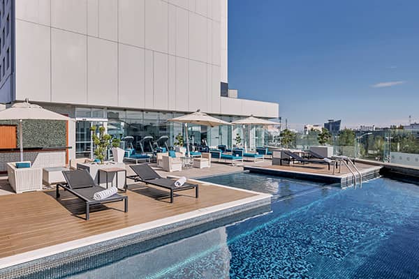 Outdoor rooftop pool area at one of Mexico’s top resorts, featuring lounge chairs, umbrellas, and glass walls. Rolled towels await guests after meetings or conference events. A small hot tub and gym are also visible under the sunny blue sky.