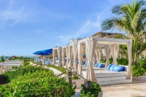 Cabañas al aire libre con cortinas blancas y cojines azules bordean un sendero rodeado de exuberante vegetación en uno de los mejores resorts de México, perfecto para eventos o reuniones junto a la piscina bajo un cielo azul brillante y palmeras que se mecen con la brisa.