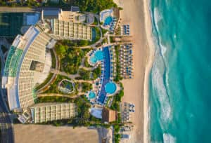 Vista aérea de un complejo turístico frente al mar en México con hoteles modernos, varias piscinas, palmeras y exuberantes jardines verdes, perfecto para eventos y reuniones junto a playas de arena y olas turquesas.