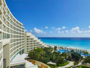 Un moderno hotel frente al mar en México, con balcones, ofrece vistas a un océano turquesa, una playa de arena, palmeras y una zona de piscina bajo un cielo azul despejado; ideal para reuniones o eventos de conferencias en el paraíso.