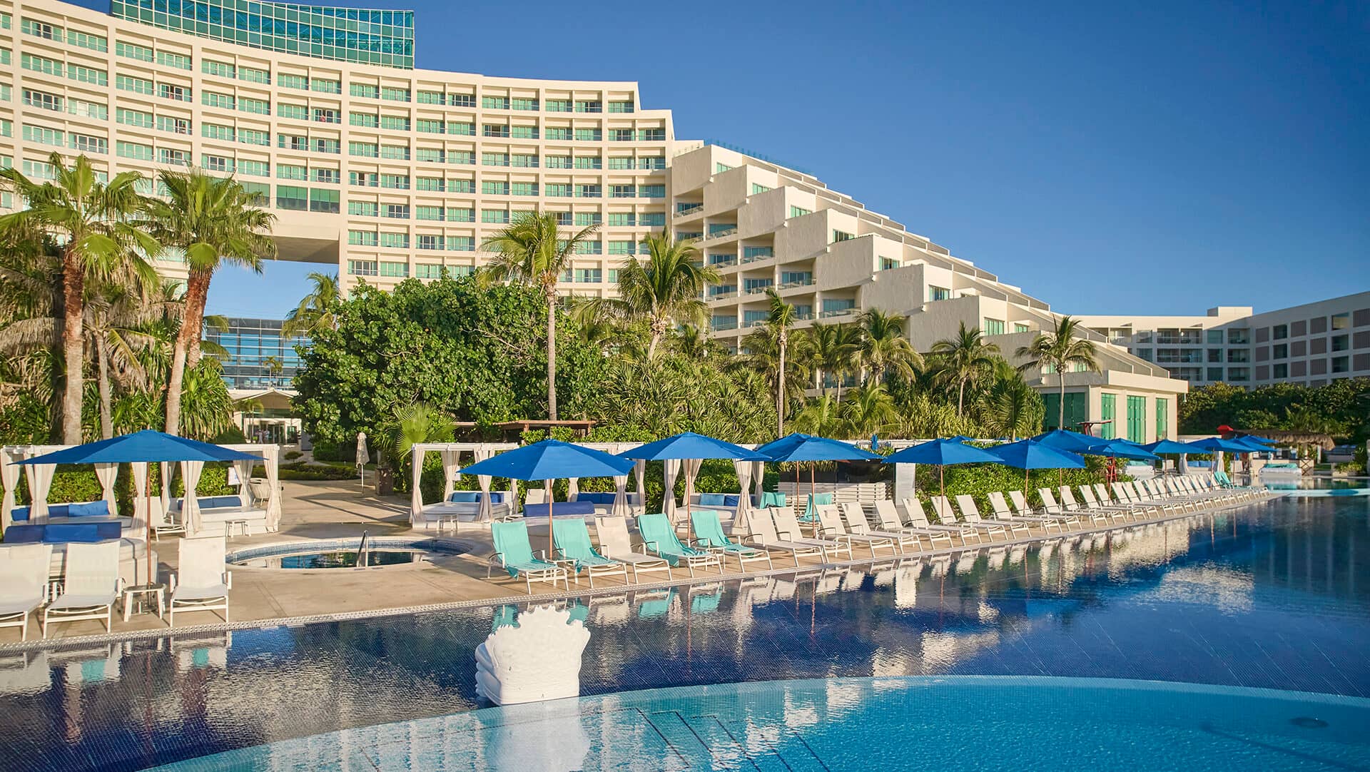 Un complejo turístico moderno con un edificio curvo, palmeras y una gran piscina al aire libre rodeada de tumbonas blancas y sombrillas azules, perfecto para reuniones o eventos bajo el cielo azul despejado.