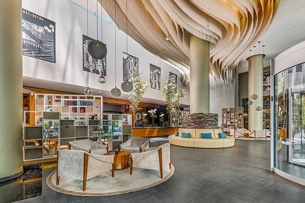 Modern hotel lobby with contemporary furniture, artistic ceiling design, large pillars, hanging lights, bookshelves, indoor trees, and black-and-white artwork. Bright and spacious atmosphere—ideal for meetings or conference events in upscale Mexico hotels.