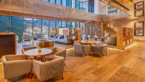 Salón moderno con suelos de madera, mobiliario en tonos neutros, mesas redondas y estanterías, ideal para reuniones o conferencias. Sus amplios ventanales ofrecen vistas panorámicas de la ciudad y las montañas. Una escalera conduce a una zona de estar adicional en este acogedor espacio inspirado en los hoteles mexicanos.