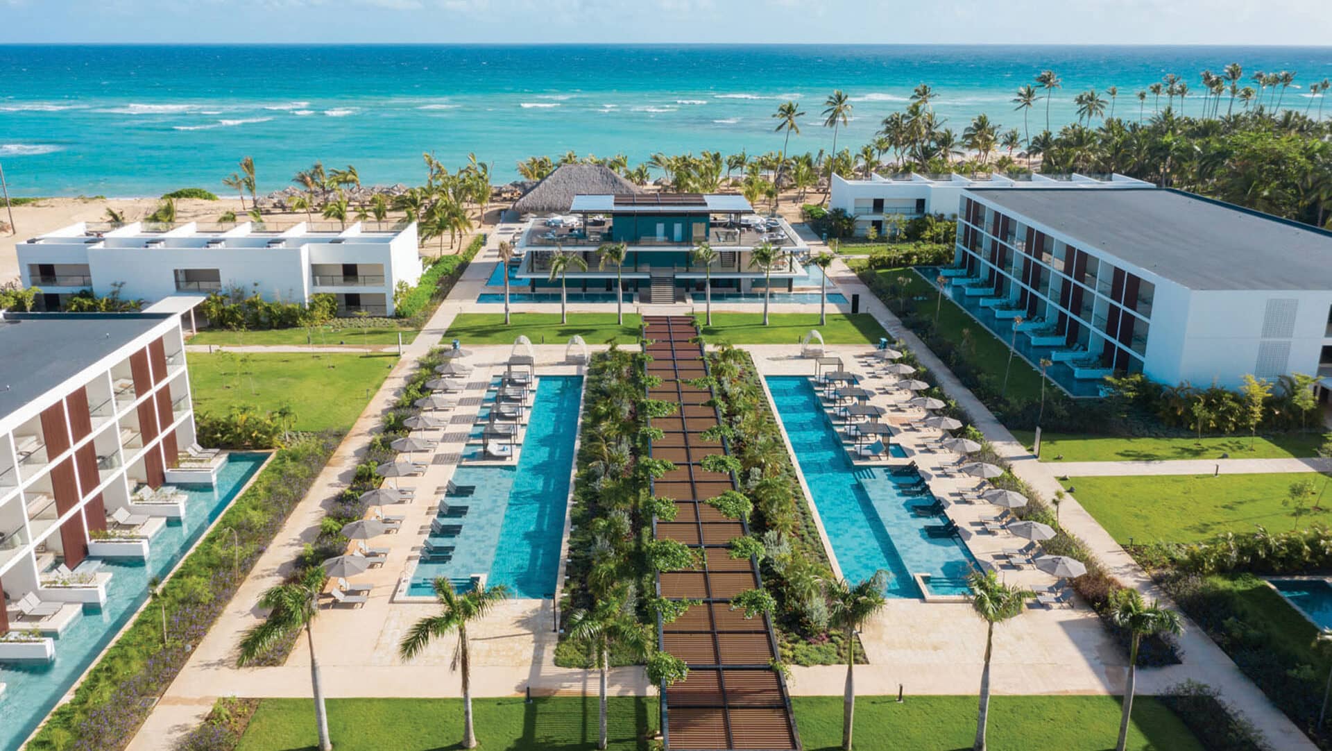 Vista aérea de un lujoso complejo turístico de playa en México con modernos edificios blancos, dos largas piscinas bordeadas de palmeras y tumbonas, exuberantes céspedes verdes y el océano azul: perfecto para reuniones o eventos de conferencias.