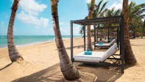 Modernas tumbonas con dosel, cojines blancos y toallas azules enrolladas bordean la playa de arena en los complejos turísticos para eventos y conferencias en México, ubicadas bajo palmeras y con vistas a las olas turquesas del océano bajo un cielo brillante y soleado.