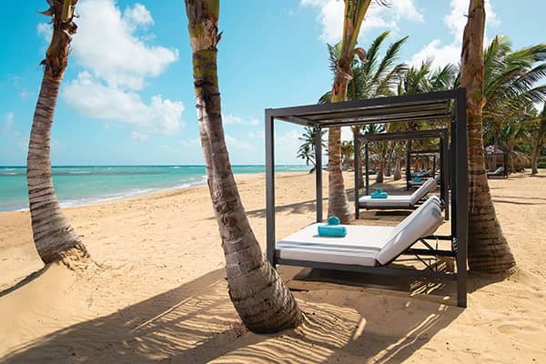 Beachside cabana beds with white cushions and blue towels sit under palm trees on a sandy shore, facing a turquoise ocean—an idyllic retreat for conference events or meetings at top Mexico resorts.