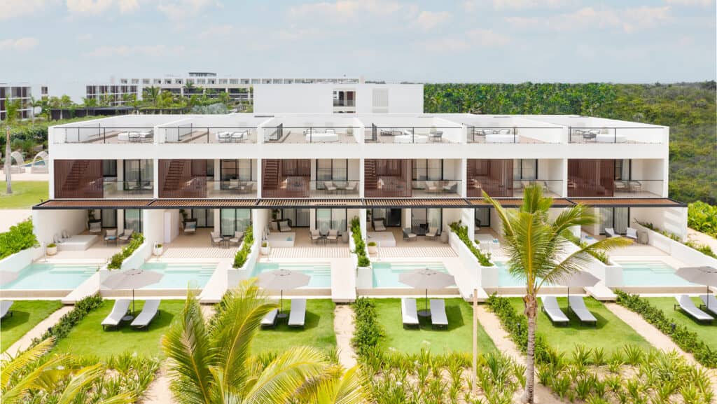 Modern white resort building with two stories, private pools, lounge chairs, and shaded patios. Perfect for conference events or meetings, this Mexico hotel features lush tropical landscaping and palm trees under a partly cloudy sky.