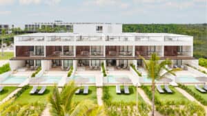 Moderno edificio blanco de dos plantas, con piscinas privadas, tumbonas y patios sombreados. Ideal para conferencias o reuniones, este hotel mexicano cuenta con exuberantes jardines tropicales y palmeras bajo un cielo parcialmente nublado.