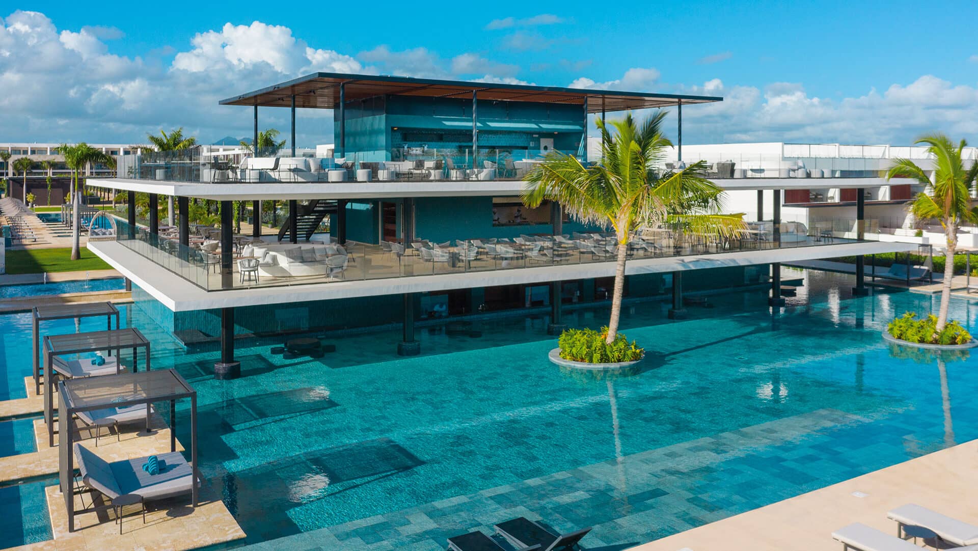 Un moderno edificio de dos plantas, ideal para conferencias y reuniones, cuenta con comedores al aire libre sobre una gran piscina de aguas cristalinas. Palmeras y cómodos sillones estilo resort rodean la piscina bajo un cielo brillante y soleado, un entorno perfecto para hoteles en México.