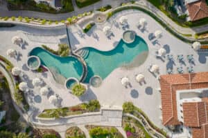Vista aérea de una piscina de forma singular con jacuzzis circulares, rodeada de tumbonas y sombrillas, ubicada en una zona ajardinada cerca de hoteles y centros de conferencias con techos de terracota en México.