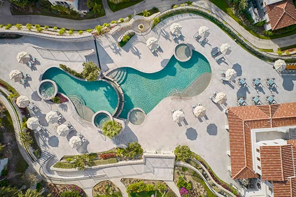 Aerial view of a curvy swimming pool with circular hot tubs at a Mexico resort, surrounded by lounge chairs and umbrellas, bordered by pathways, greenery, and terracotta-roofed buildings—ideal for conference events or relaxing after meetings.