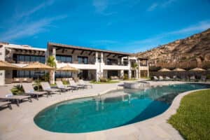 Una moderna villa de dos plantas en un complejo turístico de México, con amplios ventanales y balcones, está rodeada de tumbonas y sombrillas. Cuenta con una piscina curva y un jacuzzi, perfectos para relajarse después de conferencias o reuniones, con vistas a las colinas bajo un cielo azul despejado.