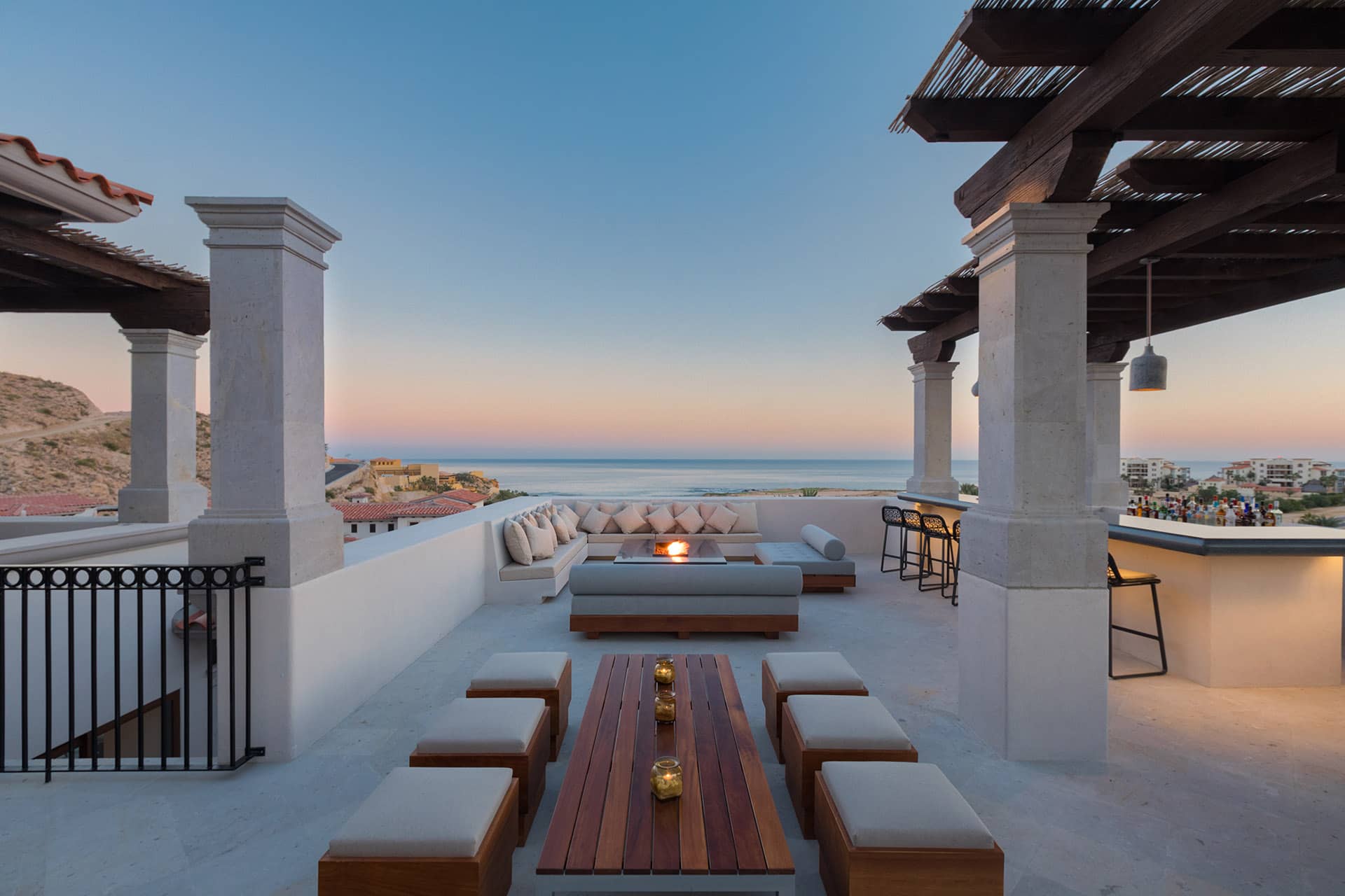 A stylish outdoor terrace at Mexico resorts features cushioned seating, a wooden table, and bar stools overlooking the ocean at sunset—an ideal setting for conference events or meetings, framed by white pillars and a dark wooden pergola with candles arranged on the table.