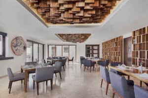 Interior de restaurante moderno con sillas tapizadas en gris y mesas de madera, ideal para reuniones o eventos. Los paneles de madera artísticos en el techo y las estanterías en las paredes realzan el ambiente, mientras que los grandes ventanales inundan este espacio de hotel en México con luz natural.