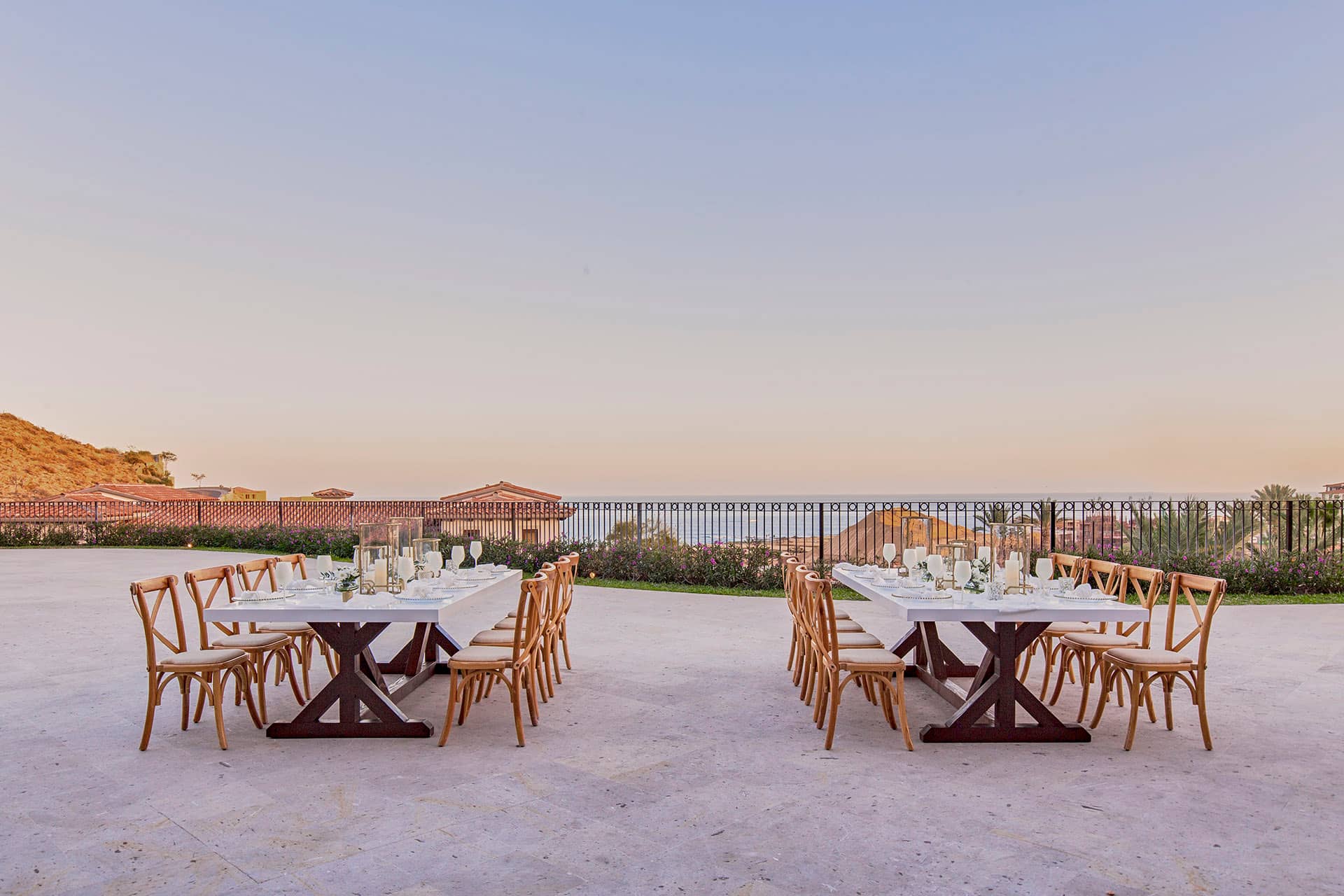 Two long dining tables with wooden chairs are set outdoors on a patio at a Mexico resort, overlooking the ocean and rooftops—perfect for conference events or meetings—with glassware and table settings arranged neatly under a clear sky.