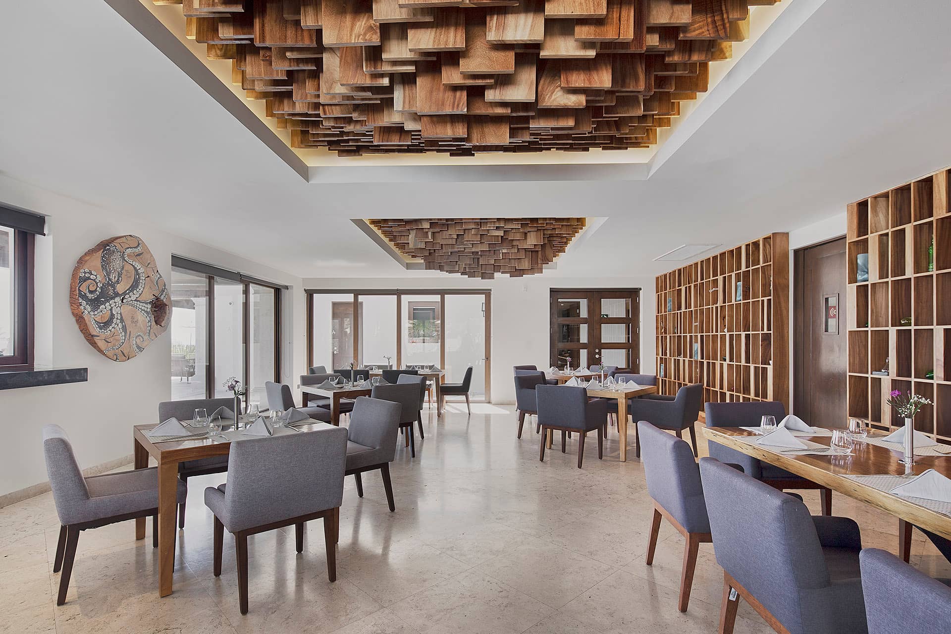 Modern restaurant interior with gray upholstered chairs and wooden tables, ideal for meetings or conference events. Artistic wooden ceiling panels and wall shelves enhance the ambiance, while large windows fill this Mexico hotel space with natural light.