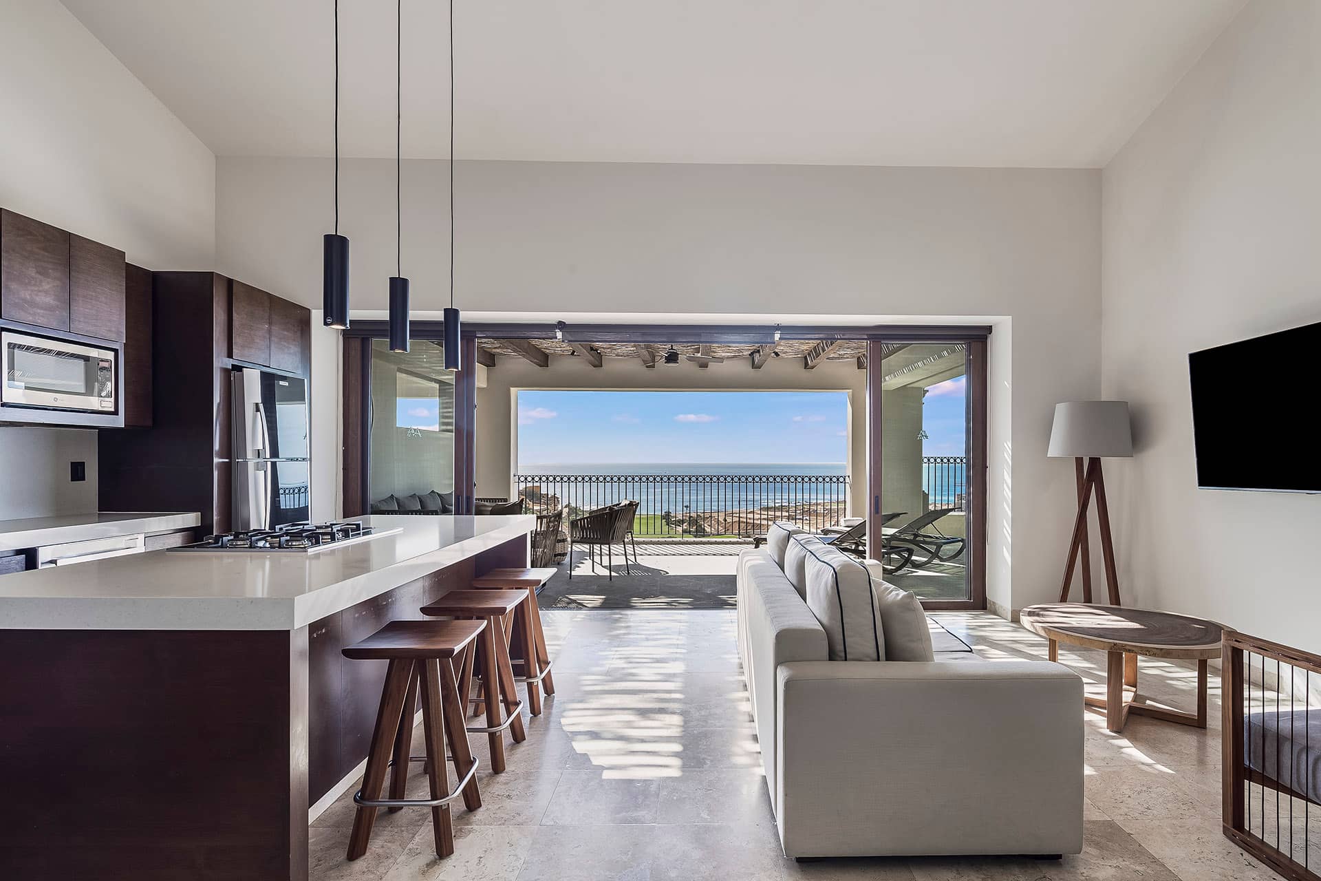 Modern kitchen and living area with a white island, wooden stools, and sofa, opening to a balcony with ocean views—a perfect retreat for Mexico hotels or conference events. Large glass doors let in abundant natural light.