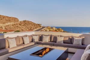 Un moderno patio al aire libre en los resorts de México, con un gran sofá modular gris, cojines estampados y una mesa rectangular con fogata, con vistas a las colinas, los tejados y el océano bajo un cielo despejado: perfecto para relajarse después de las reuniones.