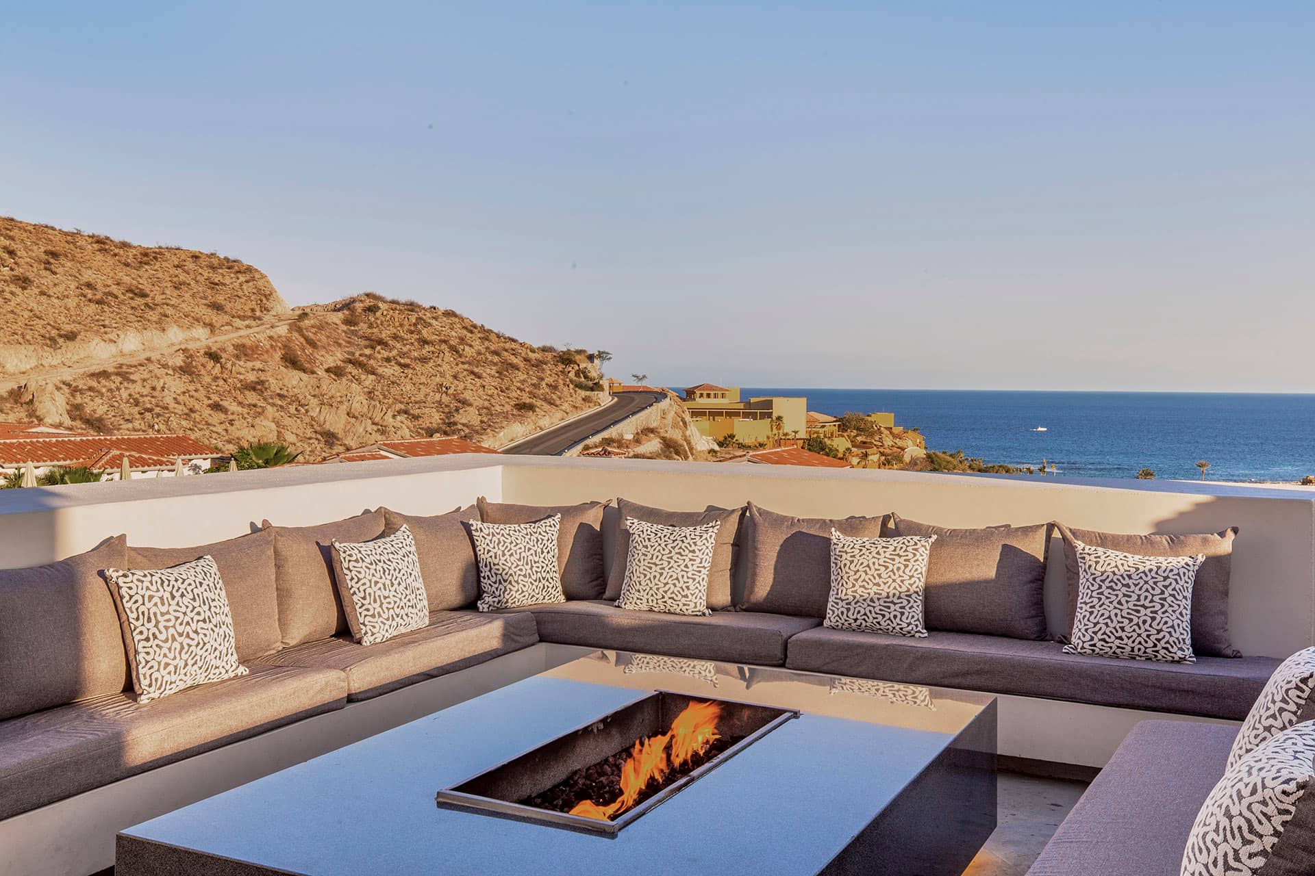 A modern outdoor patio at Mexico resorts with a large gray sectional sofa, patterned cushions, and a rectangular fire pit table, overlooking hills, rooftops, and an ocean view under a clear sky—perfect for relaxing after meetings.