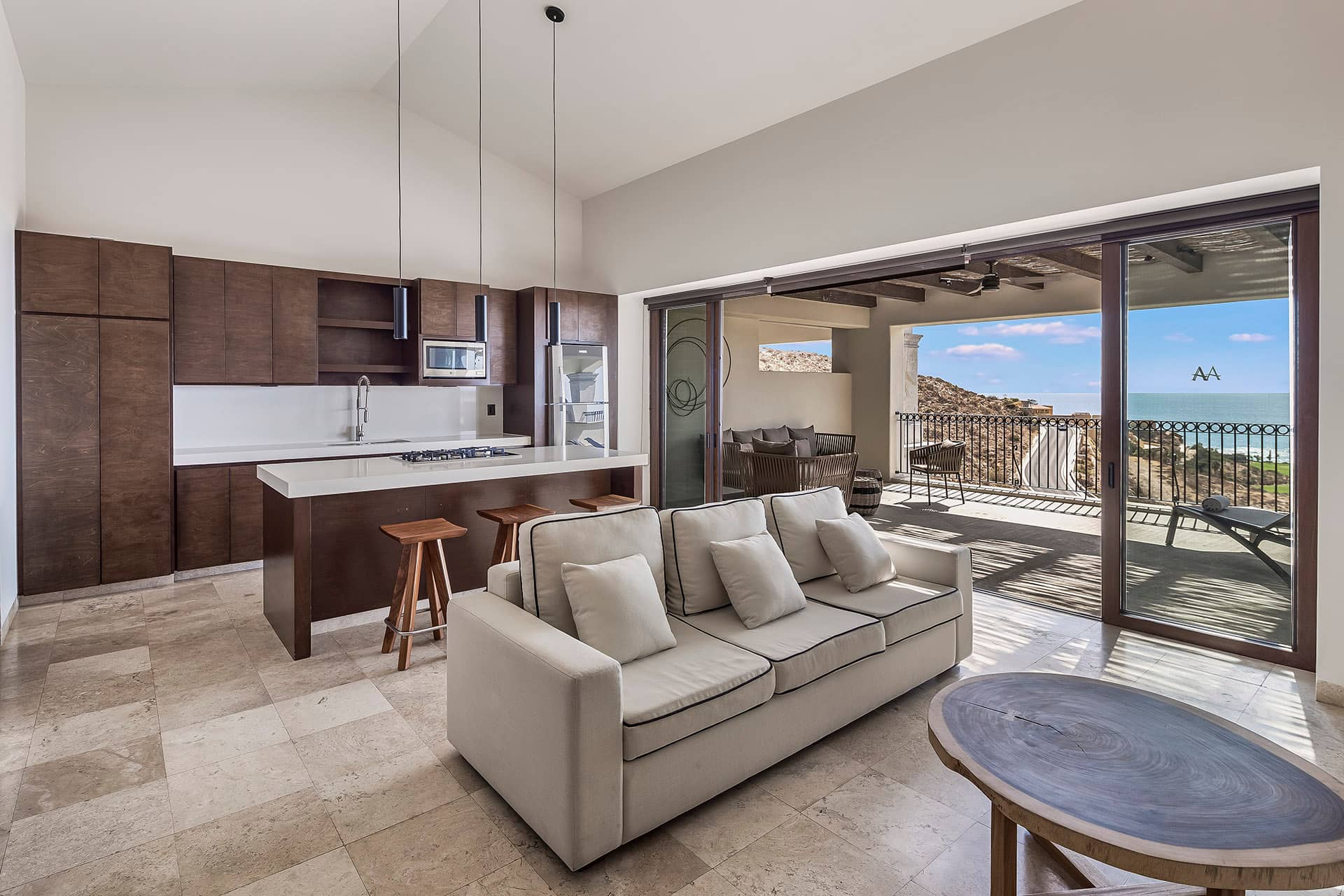 Modern open-concept kitchen and living room with brown cabinetry, a white island with stools, beige sofa, round coffee table, and large sliding doors leading to a balcony with ocean views—perfect for relaxing after meetings at Mexico resorts.