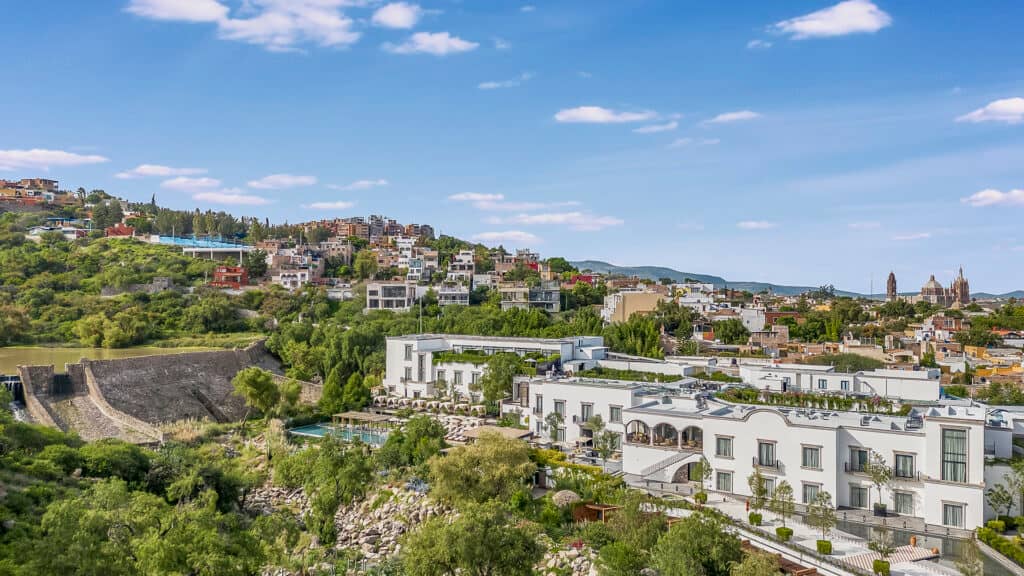 A scenic view of a hillside town with lush greenery, modern white buildings in the foreground—perfect for meetings—while colorful houses and historic architecture rise in the background under a bright blue sky with scattered clouds.