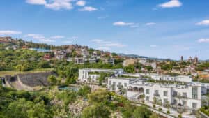 Una vista panorámica de un pueblo en la ladera de una colina con exuberante vegetación, modernos edificios blancos en primer plano —perfectos para reuniones— mientras que casas coloridas y arquitectura histórica se alzan al fondo bajo un cielo azul brillante con nubes dispersas.
