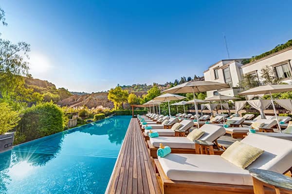 Outdoor swimming pool surrounded by wooden lounge chairs with umbrellas, rolled towels, and cushions. Modern conference events resort building and lush greenery in the background. Clear blue sky, bright sunlight, and a calm, relaxing atmosphere.