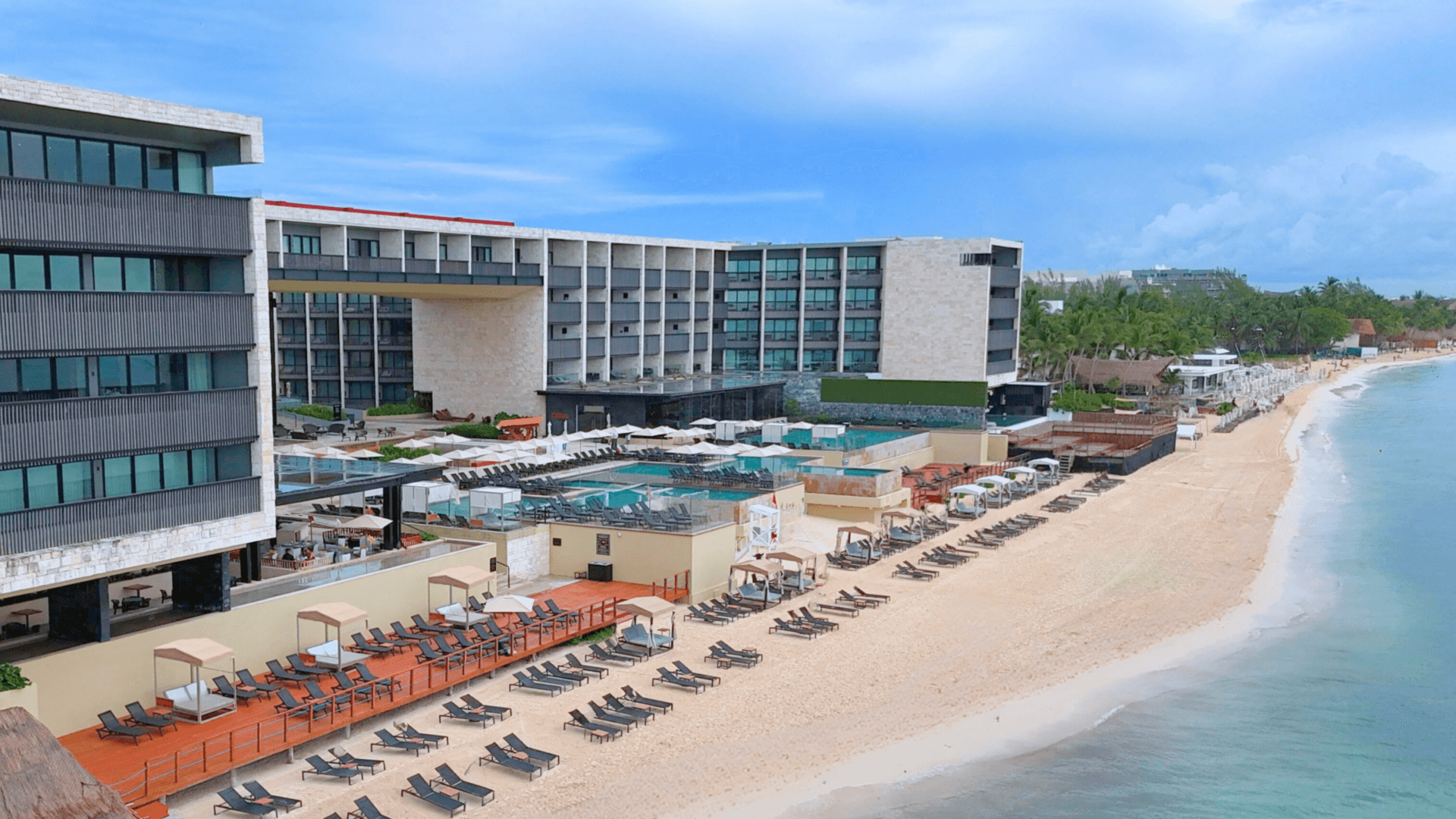 Moderno complejo turístico frente al mar con varias piscinas, tumbonas y sombrillas a lo largo de una playa de arena. Ideal para reuniones y eventos, el complejo cuenta con amplios edificios, palmeras y vistas a las cristalinas aguas azules del océano bajo un cielo parcialmente nublado.