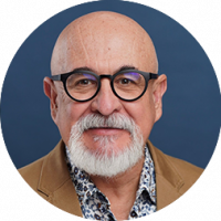 Portrait of an older man with a bald head, white beard, and mustache, wearing black-rimmed glasses, a tan blazer, and a patterned shirt—perfect for conference events or meetings at Mexico hotels—set against a solid blue background.