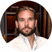 A man with long brown hair and a beard, wearing a light-colored suit jacket and a patterned shirt, poses in front of shelves with dark wood and soft lighting at one of Mexico’s premier conference resorts.