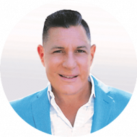 A man with short dark hair, wearing a light blue blazer over a white shirt, smiles at the camera against a light, blurred background—capturing the welcoming atmosphere of Mexico hotels known for hosting memorable conference events.
