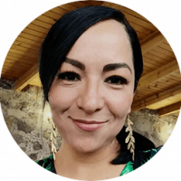 A woman with long dark hair, wearing gold leaf-shaped earrings and a green top, smiles at the camera. She is indoors at a Mexico resort, with a wooden ceiling and stone wall in the background.